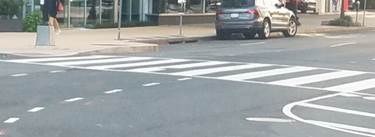 These two photos both show a crosswalk that is marked not only by a pair of white transverse lines that define the outside edges of the crosswalk, but also by a series of wide white longitudinal lines parallel to traffic flow between the transverse lines to make the crosswalk more visible to motorists.