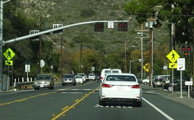 The photo shows a mast arm extending in a cantilever fashion over the crosswalk from a support pole on the left-hand side of the roadway adjacent to the crosswalk.  Mounted on the mast arm are two pedestrian hybrid beacon faces facing the camera and two pedestrian hybrid beacon faces facing the opposing direction.  None of the pedestrian hybrid beacon indications in the two pedestrian hybrid beacon faces facing the camera are illuminated.  Mounted on the mast arm between the two pedestrian hybrid beacon faces facing the camera is a CROSSWALK STOP ON RED (symbolic circular red) (R10-23) sign.  Post-mounted on the left-hand side and on the right-hand side of the roadway adjacent to the crosswalk are Pedestrian (W11-2) warning signs with a diagonal downward pointing arrow (W16-7P) plaques.  A STOP HERE ON RED (R10-6) sign is post-mounted on the right-hand side of the roadway prior to the crosswalk.