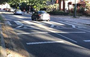 This photo shows a two-lane one-way street upon which motor vehicles are traveling away from the camera.  A marked crosswalk is shown across the roadway on a raised portion of roadway, also known as an extended speed hump or a speed table.  The standard speed hump advance markings shown in Figure 3B-31 of the 2009 MUTCD are present in both lanes.