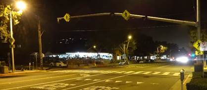 This photo shows an uncontrolled marked crosswalk.  A Yield Here To Pedestrians (R1-5a) sign is post-mounted prior to the crosswalk on the right-hand side of the roadway.  A mast arm extends in a cantilever fashion over the crosswalk from a support pole on the right-hand side of the roadway adjacent to the crosswalk.  A Pedestrian (W11-2) warning sign with a diagonal downward pointing arrow (W16-7P) plaque is mounted on the mast arm support pole.  A Pedestrian (W11-2) warning sign with a diagonal downward pointing arrow (W16-7P) plaque is mounted on a street light support pole on the left-hand side of the roadway.  Two single-section warning beacons are mounted on the mast arm for each direction of travel and neither of the yellow indications facing the camera are illuminated.  Two Pedestrian (W11-2) warning signs are mounted on the mast arm, one of which is between the two warning beacons facing the opposing direction of travel and one of which is between the two warning beacons facing the camera.  A street light (luminaire) is shown on the left-hand side of the roadway and it is providing ambient light in the crosswalk location.